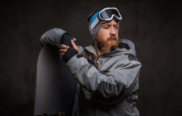 Roodharige bebaarde man met een volledige uitrusting voor extreem snowboarden, leunend op een snowboard en wegkijkend