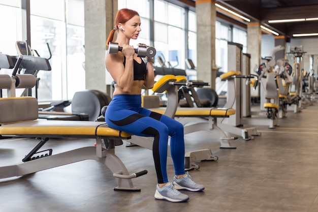 Roodharige fitness vrouw zittend op een bankje met halters in de sportschool