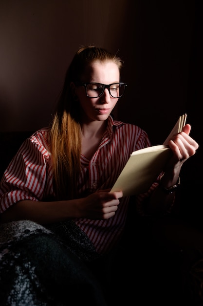 Roodharige gember meisje in glazen lezen boek, liggend op de bank