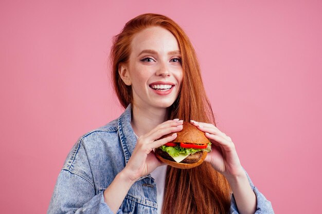 Roodharige gember vrouw met sproeten genieten van grote enorme hamburger kotelet en het dragen van demin american jeans jasje in studio roze achtergrond Usa traditionele concept