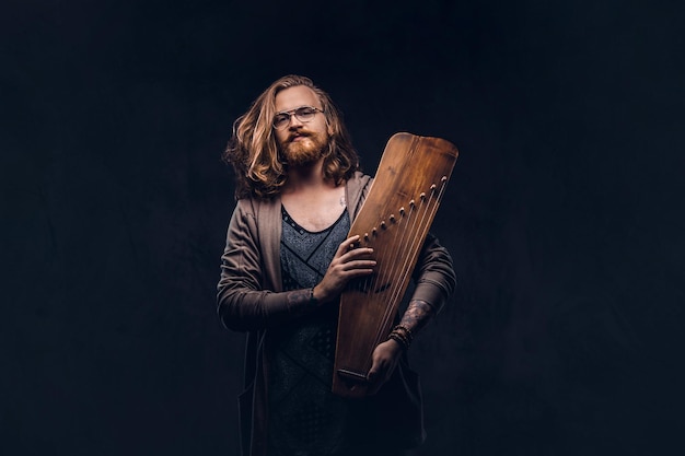 Roodharige hipster man met lang weelderig haar en volle baard gekleed in vrijetijdskleding heeft een Russisch traditioneel muziekinstrument - gusli. Geïsoleerd op een donkere achtergrond.