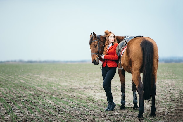 Roodharige jockeyvrouw in een rood vest en zwarte hoge laarzen met een paard voor een wandeling