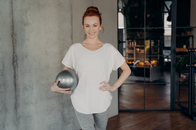 Roodharige jonge lachende vrouw met fitball in de ene hand draagt witte sport tshirt en grijze legging klaar voor barre training in fitnessstudio. Gezonde levensstijl en pilates indoor concept