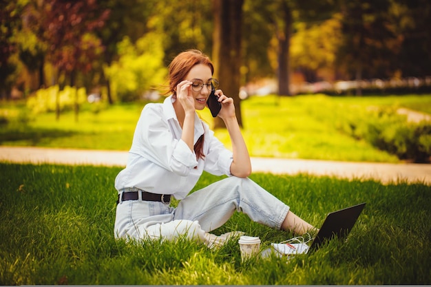 Roodharige jonge vrouw werkt buitenshuis met laptop en mobiel terwijl ze freelance op het gras zit