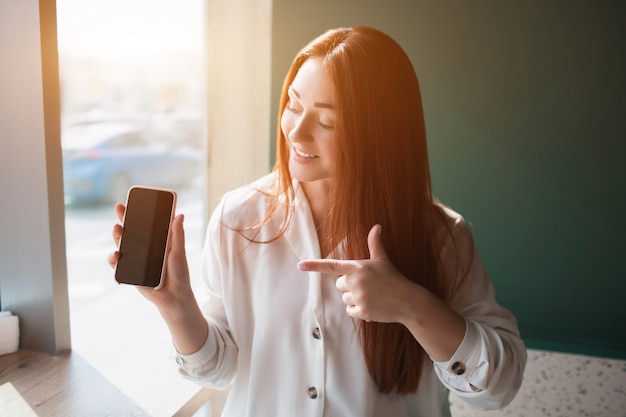 Roodharige jonge vrouw zitten in een cafe en met een telefoon. Het vrouwelijke model wijst met een wijsvinger naar de smartphone.