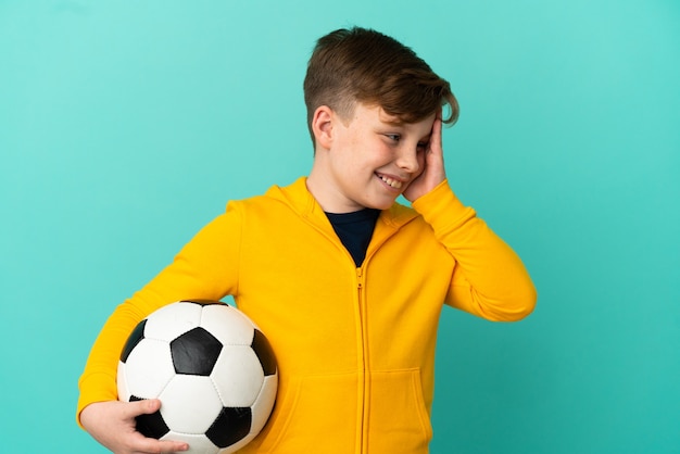 Roodharige jongen die voetbal speelt geïsoleerd op een blauwe achtergrond die veel lacht
