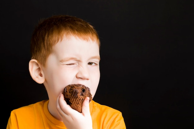 Roodharige jongen met een chocolade cupcake, een jongen eet een cupcake met plezier, zoet schadelijk maar heerlijk eten bij een kind