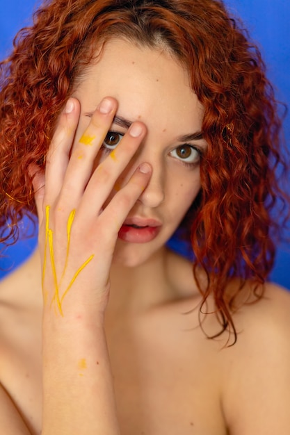 Roodharige krullende vrouw handen in gele verf kijkt naar de camera op een blauwe achtergrond. Conceptfotografie voor kunst- of vrouwenblog
