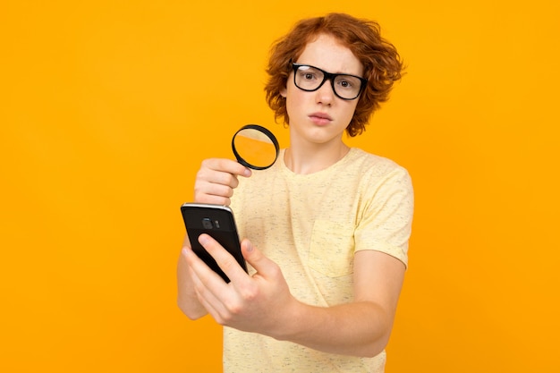 Roodharige man in een t-shirt kijkt naar een smartphone door een vergrootglas op een geel