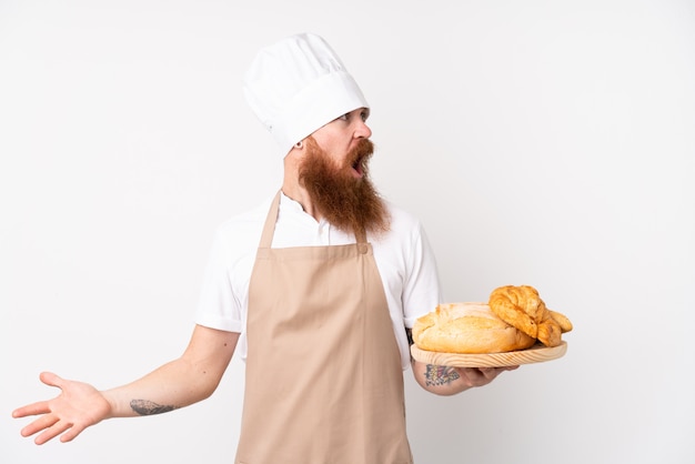 Roodharige man in uniform chef. mannelijke bakker die een lijst met verscheidene broden met verrassingsgelaatsuitdrukking houdt