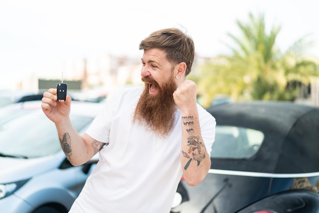 Foto roodharige man met baard die autosleutels vasthoudt terwijl hij buiten een overwinning viert