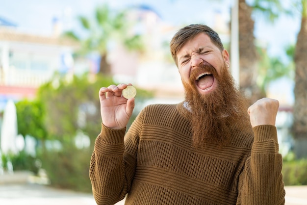 Foto roodharige man met baard die een bitcoin vasthoudt terwijl hij buiten een overwinning viert