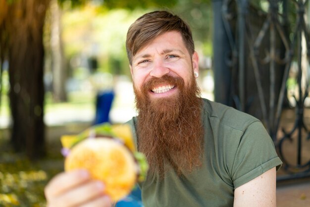 Roodharige man met baard die een hamburger in de open lucht vasthoudt met een gelukkige uitdrukking