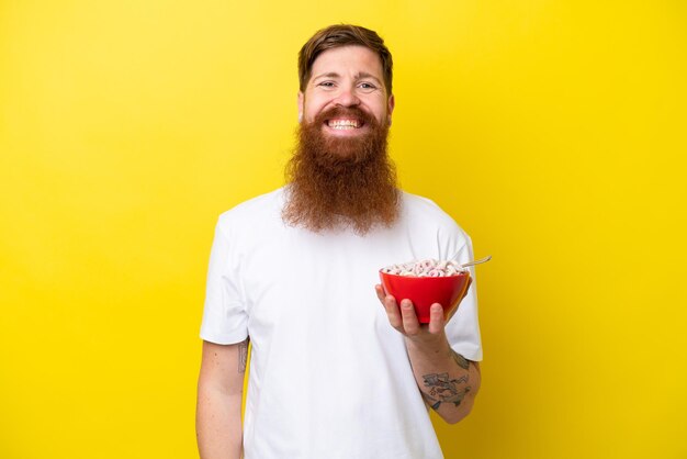 Roodharige man met baard die een kom ontbijtgranen eet geïsoleerd op een gele achtergrond die veel lacht