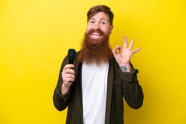 Roodharige man met baard die een microfoon oppakt die op een gele achtergrond wordt geïsoleerd en een ok teken met vingers toont