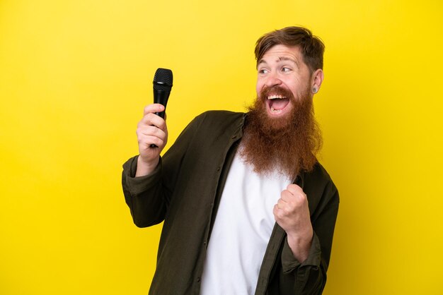 Roodharige man met baard die een microfoon oppakt die op gele achtergrond wordt geïsoleerd en een overwinning viert