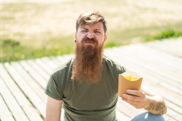 Roodharige man met baard die gebakken frites in de buitenlucht houdt met een droevige uitdrukking