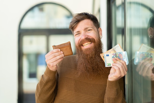 Roodharige man met baard die portemonnee met geld vasthoudt en ernaar kijkt