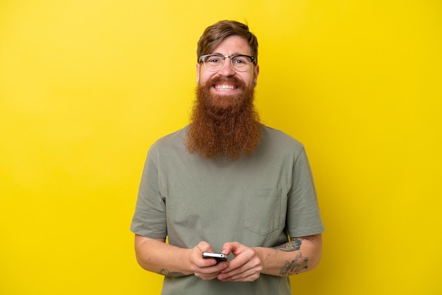 Roodharige man met baard geïsoleerd op gele achtergrond die een bericht verzendt met de mobiel