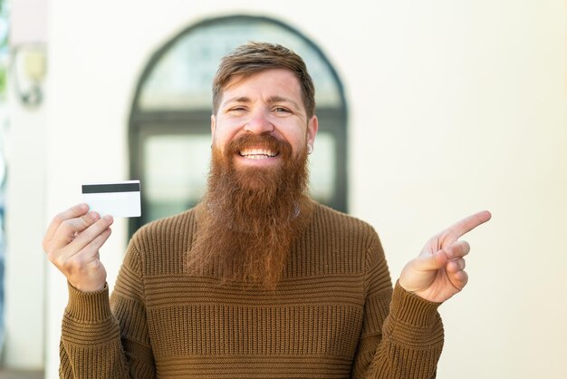 Roodharige man met baard met een creditcard in de buitenlucht verrast en wijzende vinger naar de zijkant