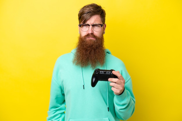 Roodharige man met baard spelen met een video game controller geïsoleerd op gele achtergrond met droevige uitdrukking