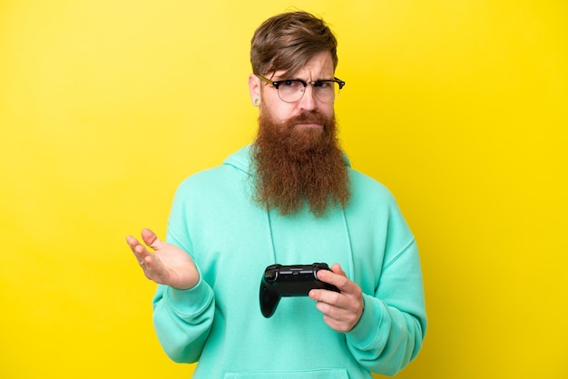 Roodharige man met baard spelen met een videogamecontroller geïsoleerd op gele achtergrond die twijfels maakt terwijl hij de schouders opheft