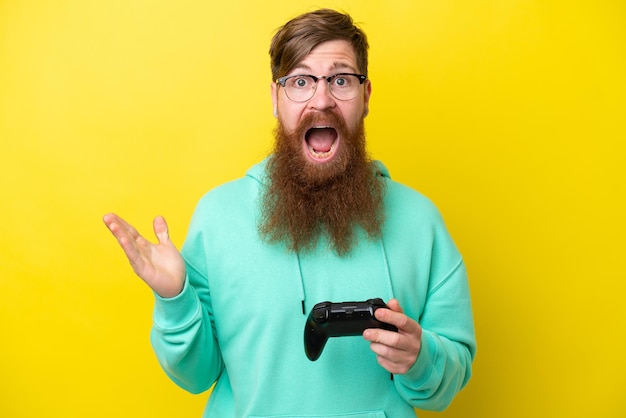 Roodharige man met baard spelen met een videogamecontroller geïsoleerd op gele achtergrond met geschokte gezichtsuitdrukking