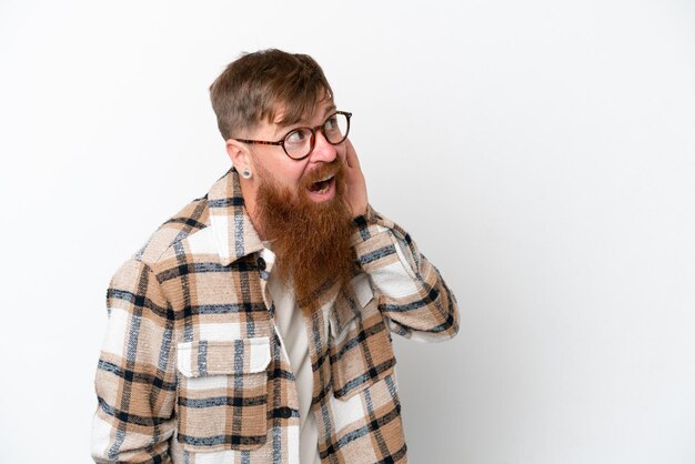 Roodharige man met lange baard geïsoleerd op witte achtergrond luisterend naar iets door de hand op het oor te leggen