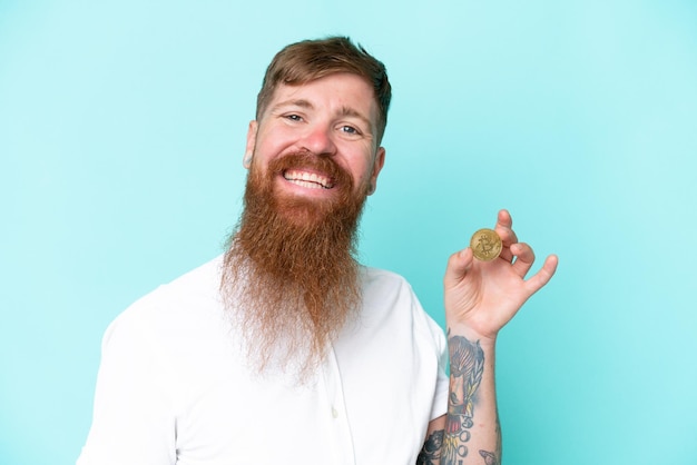 Roodharige man met lange baard met een Bitcoin geïsoleerd op een blauwe achtergrond die veel lacht