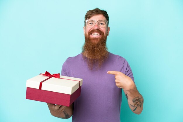 Roodharige man met lange baard met een geschenk geïsoleerd op blauwe achtergrond met verrassende gezichtsuitdrukking