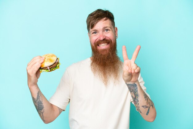 Roodharige man met lange baard met een hamburger geïsoleerd op blauwe achtergrond glimlachend en overwinningsteken tonend