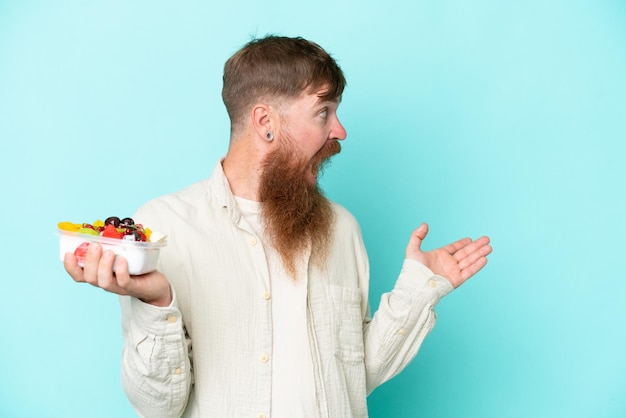 Roodharige man met lange baard met een schaal fruit geïsoleerd op blauwe achtergrond met verrassende gezichtsuitdrukking