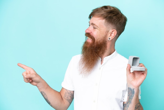 Roodharige man met lange baard met een verlovingsring geïsoleerd op blauwe achtergrond wijzend naar de zijkant om een product te presenteren