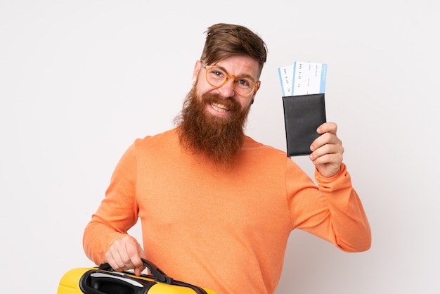 Roodharige man met lange baard over witte muur in vakantie met koffer en paspoort