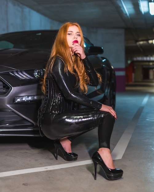 Roodharige meid in latex, een zwarte top en strakke broek met grote hakken, gehurkt in een sportwagen in een garage met een verleidelijke look