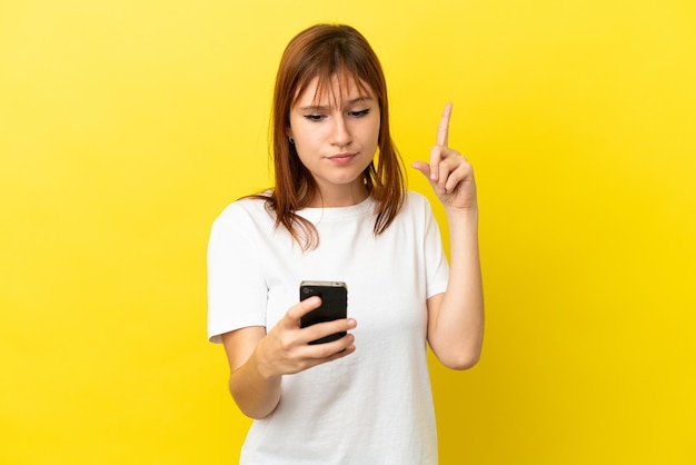 Roodharige meisje geïsoleerd op gele achtergrond met behulp van mobiele telefoon en opheffing van de vinger