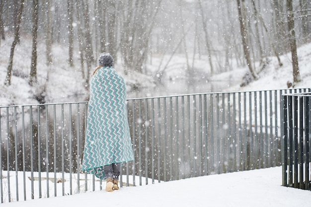 Roodharige meisje in grijze hoed, bedekt met blauwe patronen plaid genieten van sneeuwval staande achterkant op de brug in de buurt van de rivier