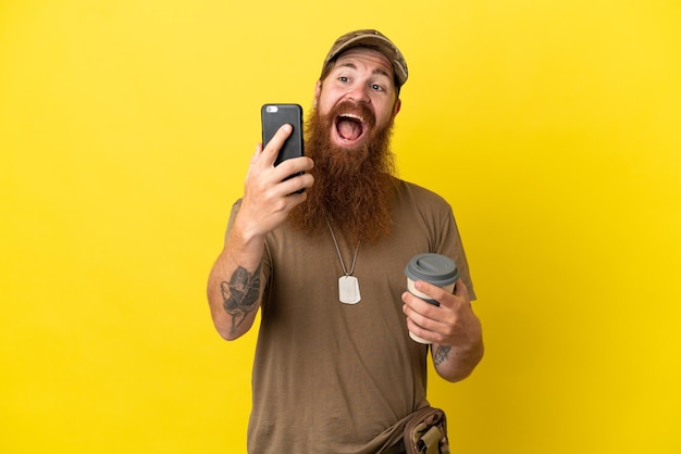 Roodharige Militaire man met dog tag geïsoleerd op gele achtergrond met koffie om mee te nemen en een mobiel