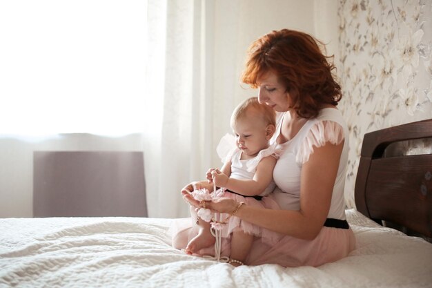 Foto roodharige moeder omhelst voorzichtig peuter baby meisje en spelen met haar op het bed in de echte casual kamer daglicht