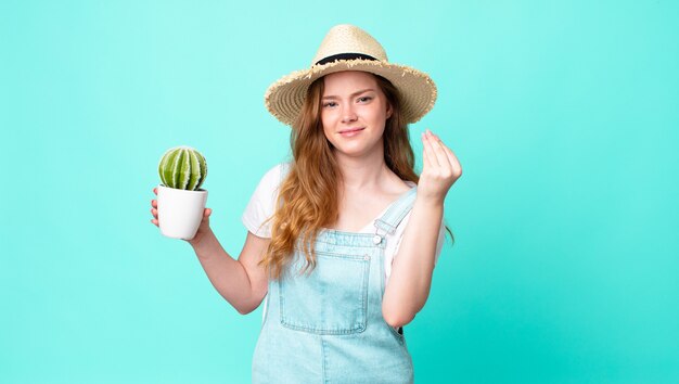 Roodharige mooie boerenvrouw die capice of geldgebaar maakt, zegt dat je moet betalen en een cactus vasthoudt