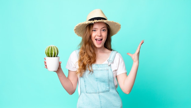 Roodharige mooie boerenvrouw die zich gelukkig voelt, verrast een oplossing of idee realiseert en een cactus vasthoudt