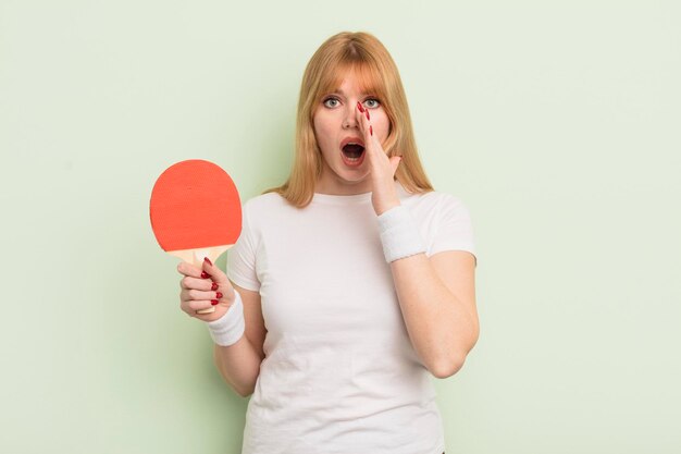 Roodharige mooie vrouw die zich gelukkig voelt en een grote schreeuw geeft met handen naast het pingpongconcept van de mond