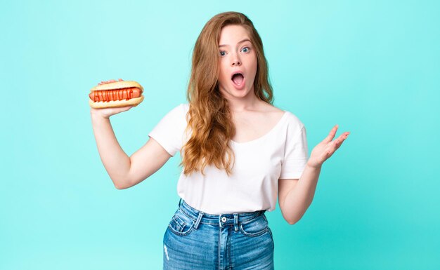 Roodharige mooie vrouw verbaasd, geschokt en verbaasd met een ongelooflijke verrassing en met een hotdog