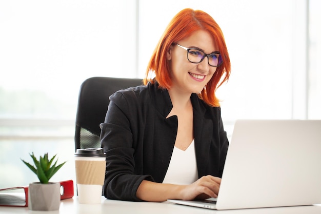 Roodharige positieve schattige mooie zakenvrouw zit binnenshuis op kantoor met behulp van een laptopcomputer