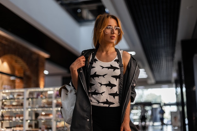 Roodharige prachtige jonge vrouw in een vintage jasje in een t-shirt met bril in een t-shirt met een patroon van stands in een moderne winkel. Modieus vrij aantrekkelijk meisjesmodel loopt op winkel.