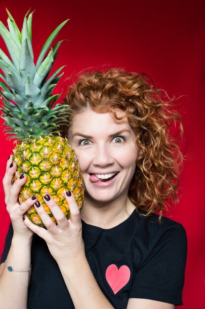 Roodharige schoonheid in een zwart t-shirt met een hartprint houdt een ananas in haar handen op een rode ruimte