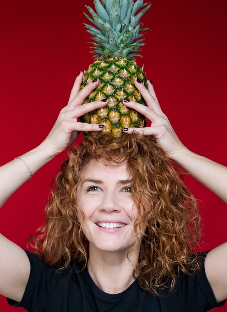 Roodharige schoonheid in een zwart t-shirt met een hartprint houdt een ananas in haar handen op een rode ruimte