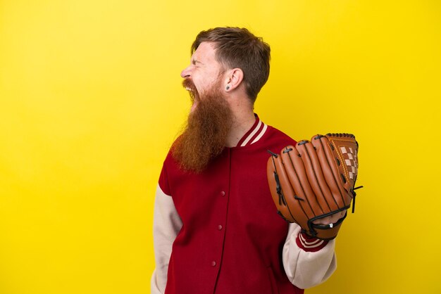 Foto roodharige speler man met baard met honkbalhandschoen geïsoleerd op gele achtergrond lachend in laterale positie