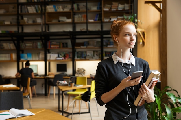 Roodharige student poseren binnenshuis in bibliotheek met boeken istening muziek met oortelefoons met behulp van mobiele telefoon