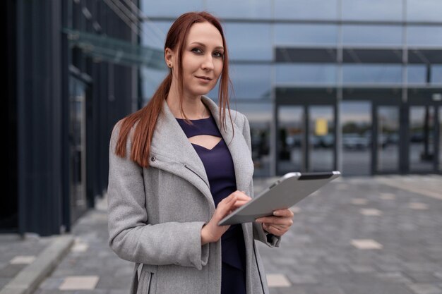 Roodharige veertigjarige zakenvrouw tegen de achtergrond van een kantoorgebouw met een tablet erin